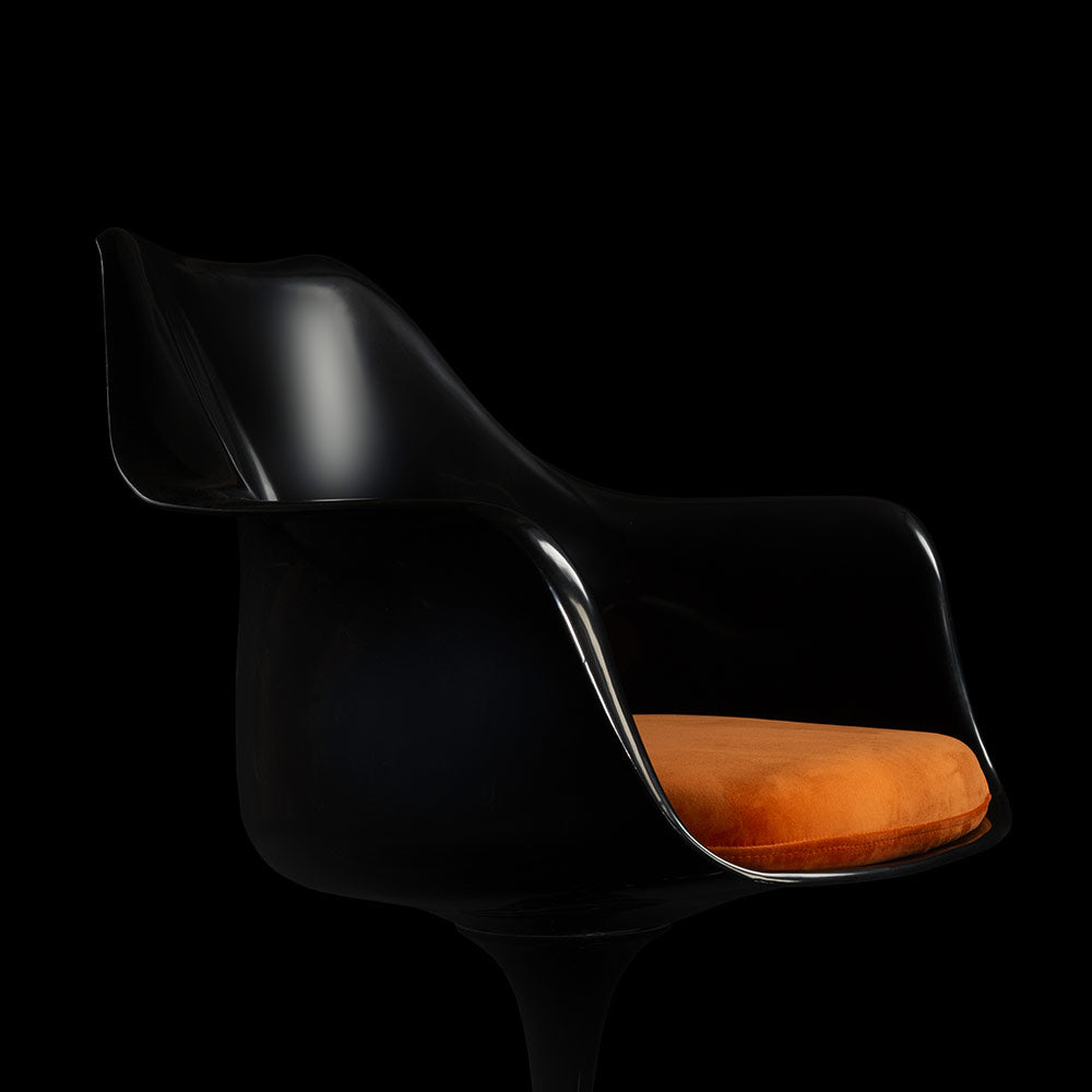 A streak of light shimmers down upon a dark and black coloured Saarinen Tulip Arm Chair with the fiery orange cushion prominent against the dark backdrop