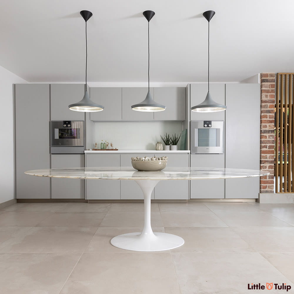 Under the lights, the grey veined Arabescato Marble 244 x 137 cm Oval Tulip Table is seen within a neutral interior setting and exposed brickwork