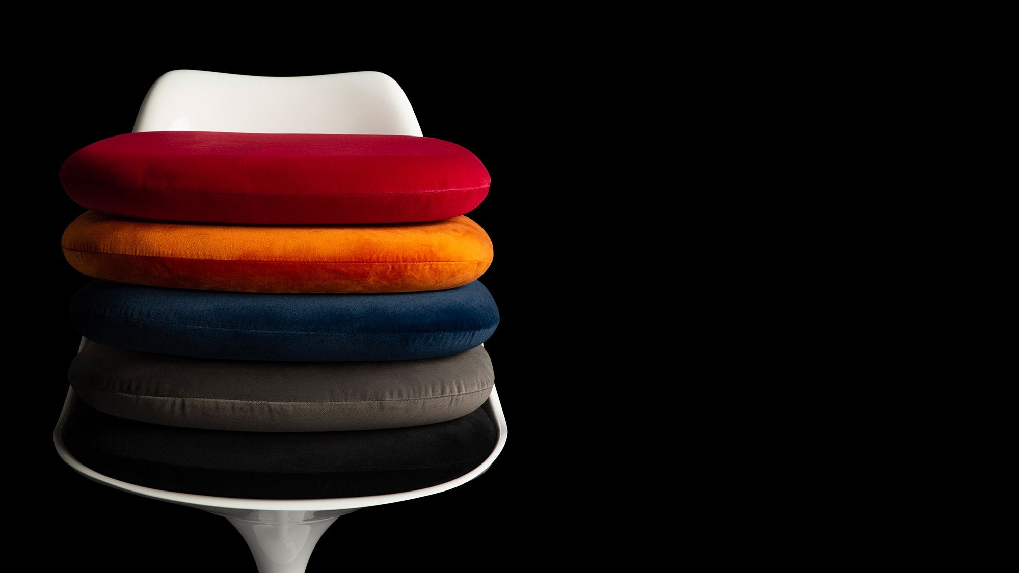 Against a black backdrop is a white Saarinen Tulip Chair face on with a stack of 5 cushion colours in red, black, blue, grey and orange