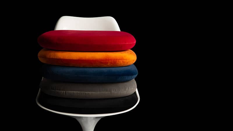 A white Tulip Chair stands in the foreground of a black background, with a tower of side cushions displaying the variety of colours and fabric available