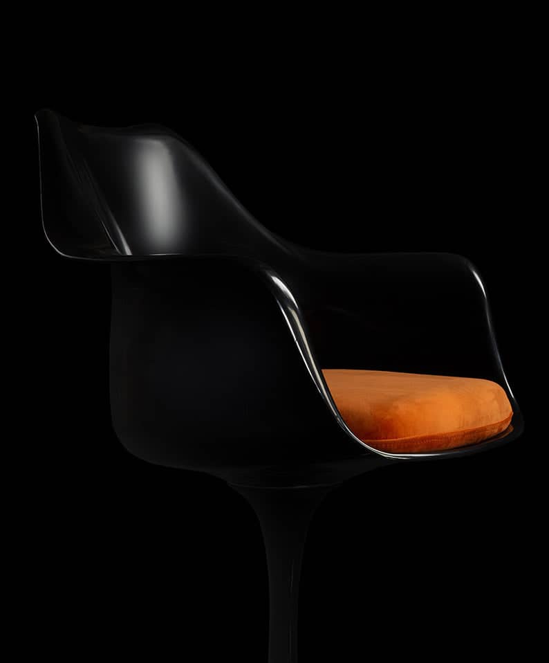 A silhouetted image of a black Saarinen Tulip Arm Chair with a contrasting orange cushion is seen lit from the top against a black dark back drop