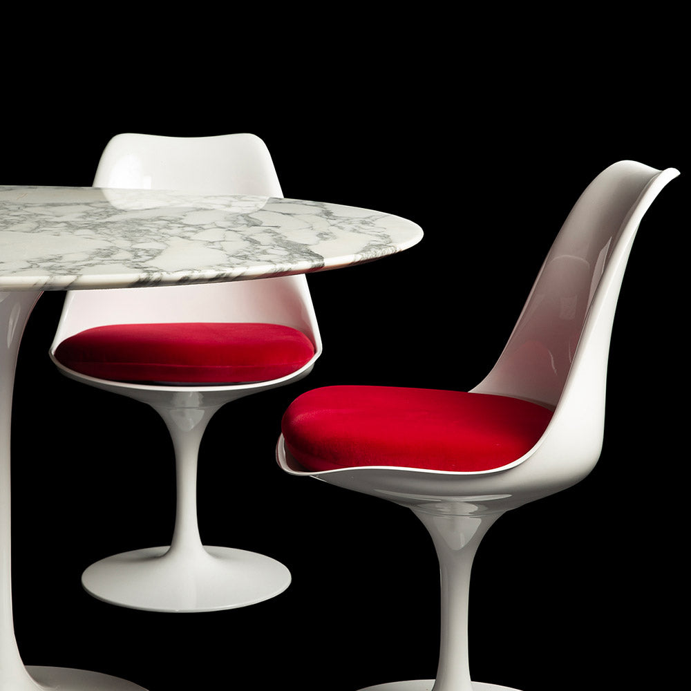 Two Tulip Side Chairs in white, one side and one front on, seen complete with deep red cushions next to a 90 cm Arabescato Tulip Table in this set image