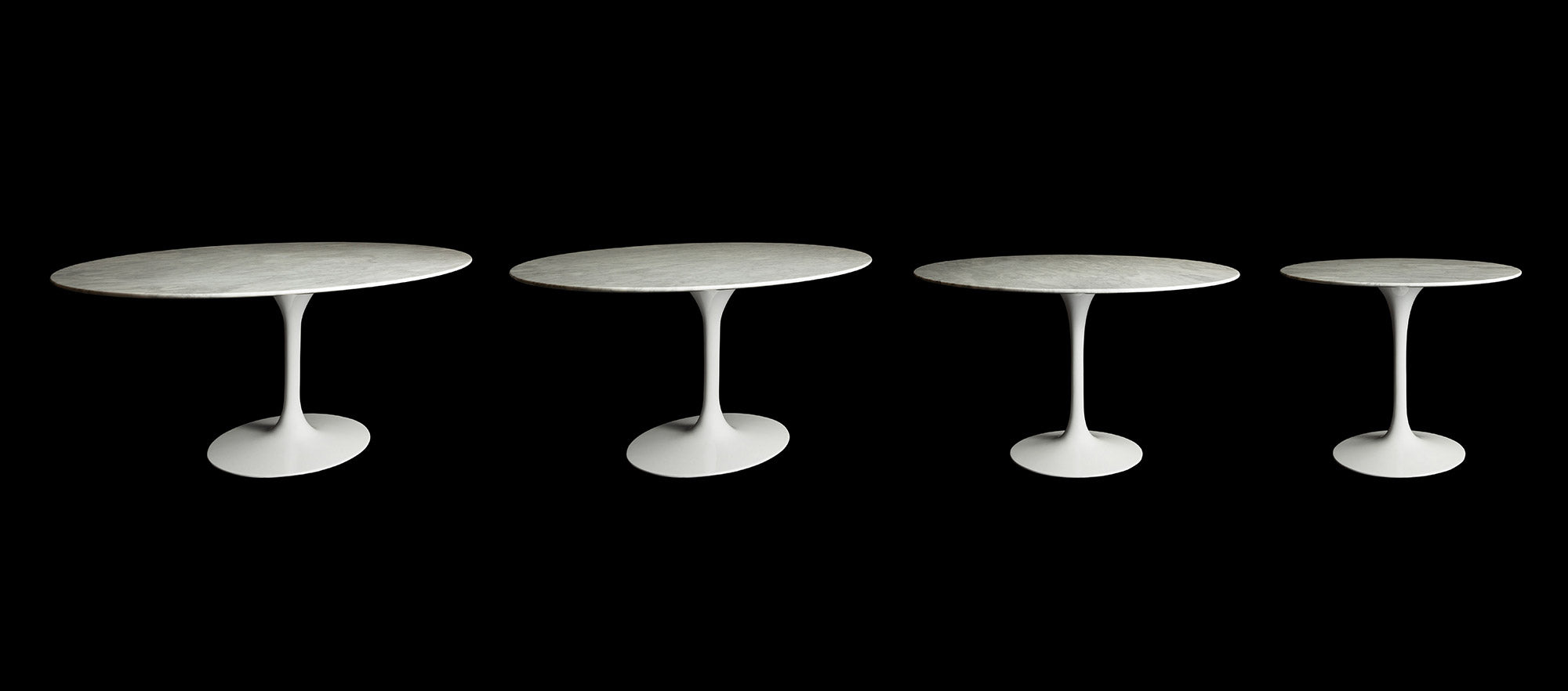 A row of Solid White Carrara Marble Tulip Tables in circular and oval shapes stand in line and stand out beautifully against the black behind them