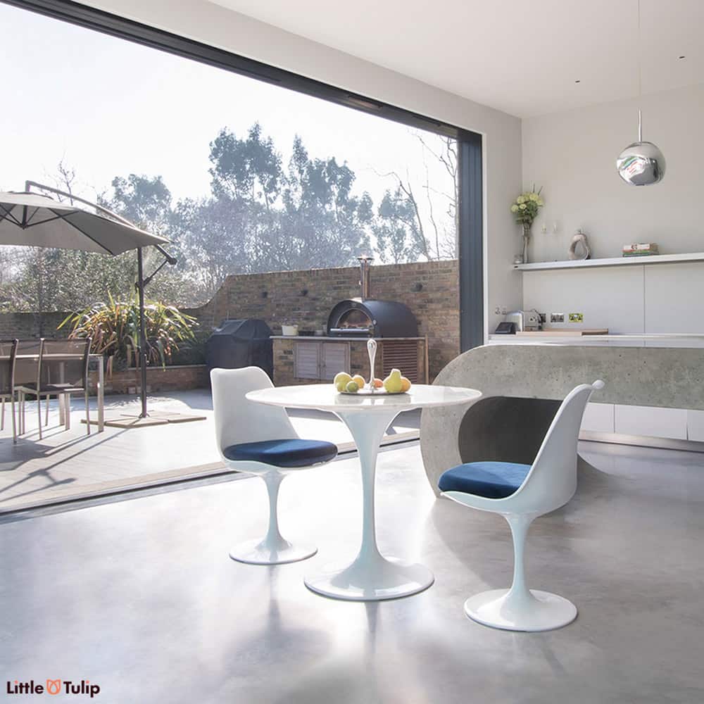An open plan kitchen with bifold doors is a backdrop to this Carrara Marble small round 90 cm Tulip Table with 2 matching side chairs with blue cushions