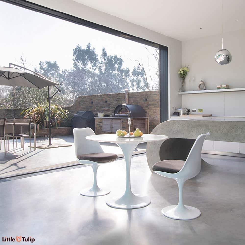 An open plan kitchen with bifold doors is a backdrop to this Carrara Marble small round 90 cm Tulip Table with 2 matching side chairs with grey cushions