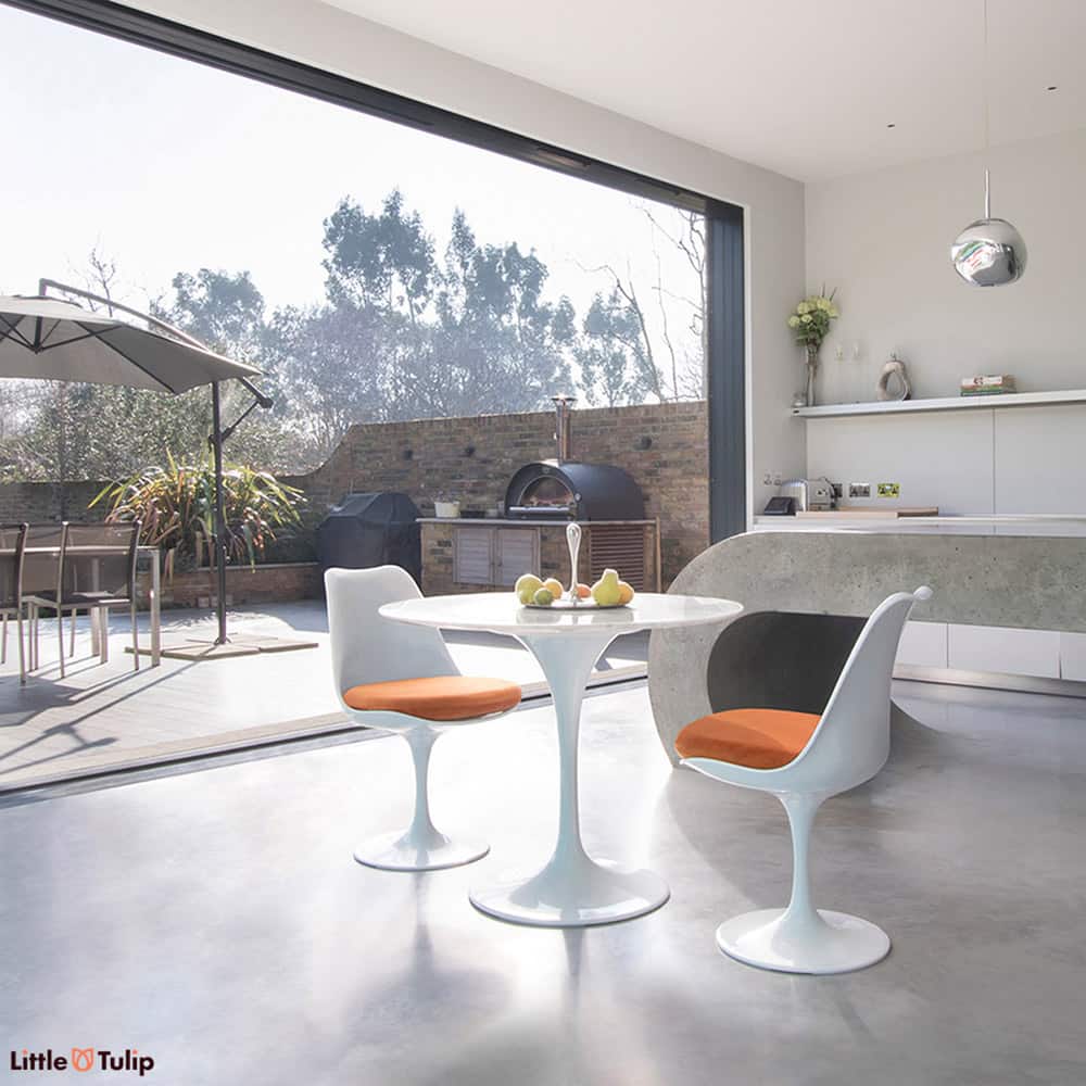 An open plan kitchen with bifold doors is a backdrop to this Carrara Marble small round 90 cm Tulip Table with 2 matching side chairs with orange cushions