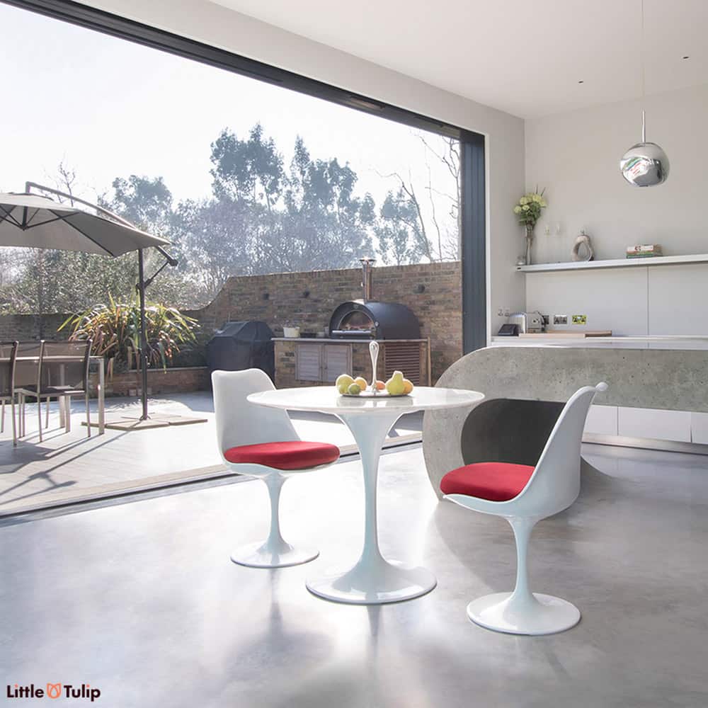 An open plan kitchen with bifold doors is a backdrop to this Carrara Marble small round 90 cm Tulip Table with 2 matching side chairs with red cushions