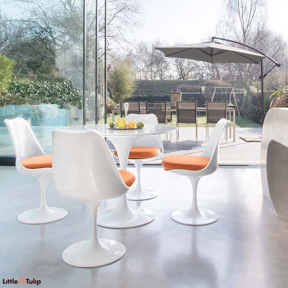 Within a modern kitchen and looking out to summer patio scene is the Carrara Marble 90cm Tulip Table with four side chairs with hot orange cushion pads